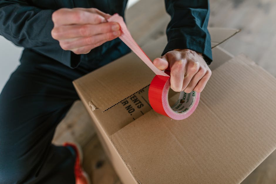 A person wearing a maroon t-shirt is sealing a large cardboard box with red packing tape, using their right hand to pull the tape dispenser and their left hand to steady the box. The individual is wea